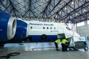 Two people wearing hi vis jacket bend over looking inside a metal unit on wheels, beside a blue and white aircraft