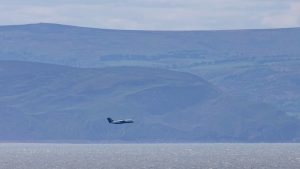 An atmospheric research aircraft flying at a low level over the sea, with hills in the background.