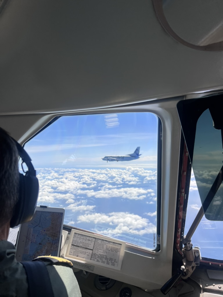 A view from an aircraft cockpit window of a white aircraft with “Cranfield” written on the fuselage flying above white clouds.