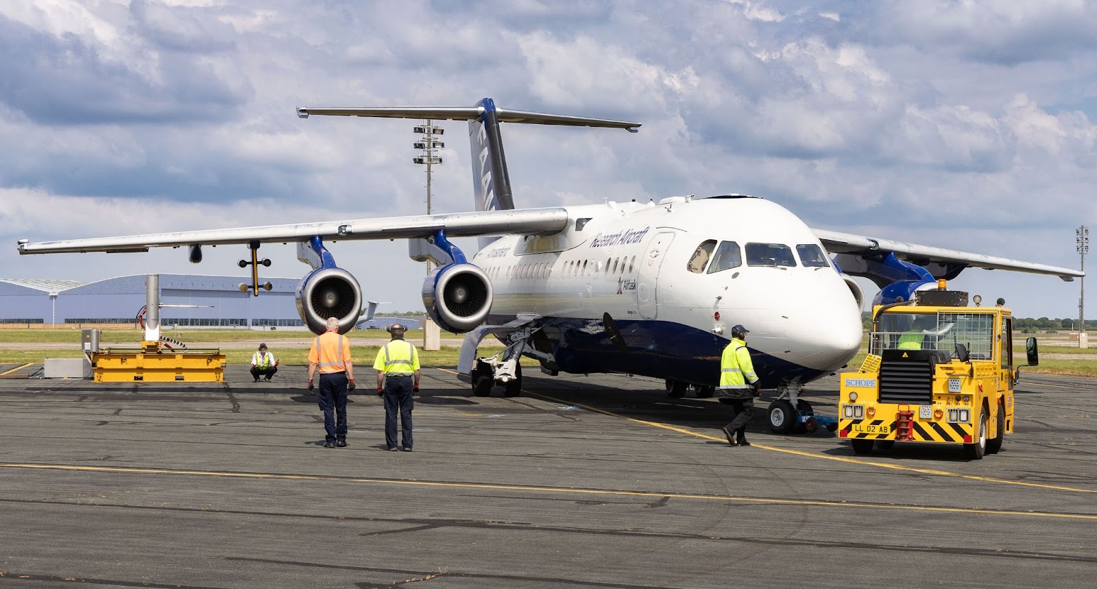 A large blue and white research aircraft being towed by a yellow aircraft tractor, with atmospheric emissions monitoring equipment behind it. Several people in hi-vis are working around the aircraft.
