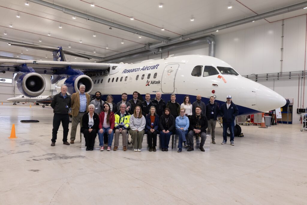 A group of people sit and stand in front of an aircraft in a hangar.
