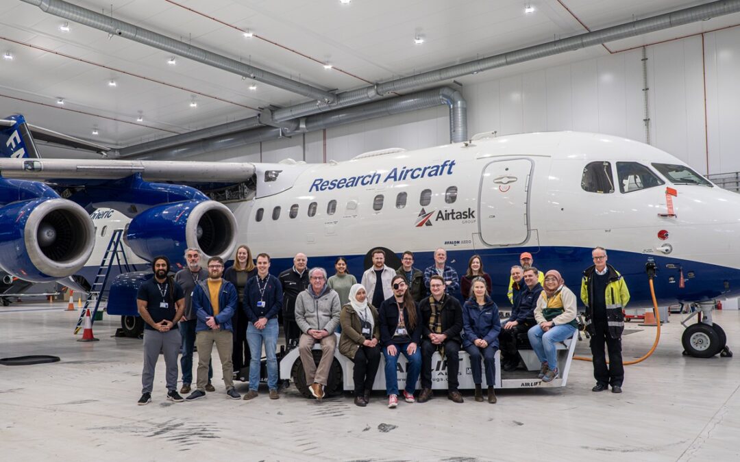 A group of people sit and stand in front of an aircraft in a hangar.